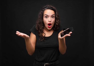 Genç ve güzel bir kadın ağzı açık ve şaşırmış, şok olmuş ve şaşırmış bir şekilde elinde bir akıllı telefon tutarak inanılmaz bir sürpriz yapıyor.