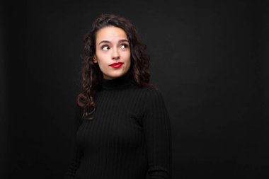 Genç ve güzel bir kadın şaşkın ve şaşkın görünüyor, merak ediyor ya da bir sorunu çözmeye çalışıyor ya da siyah arka plana karşı düşünüyor..