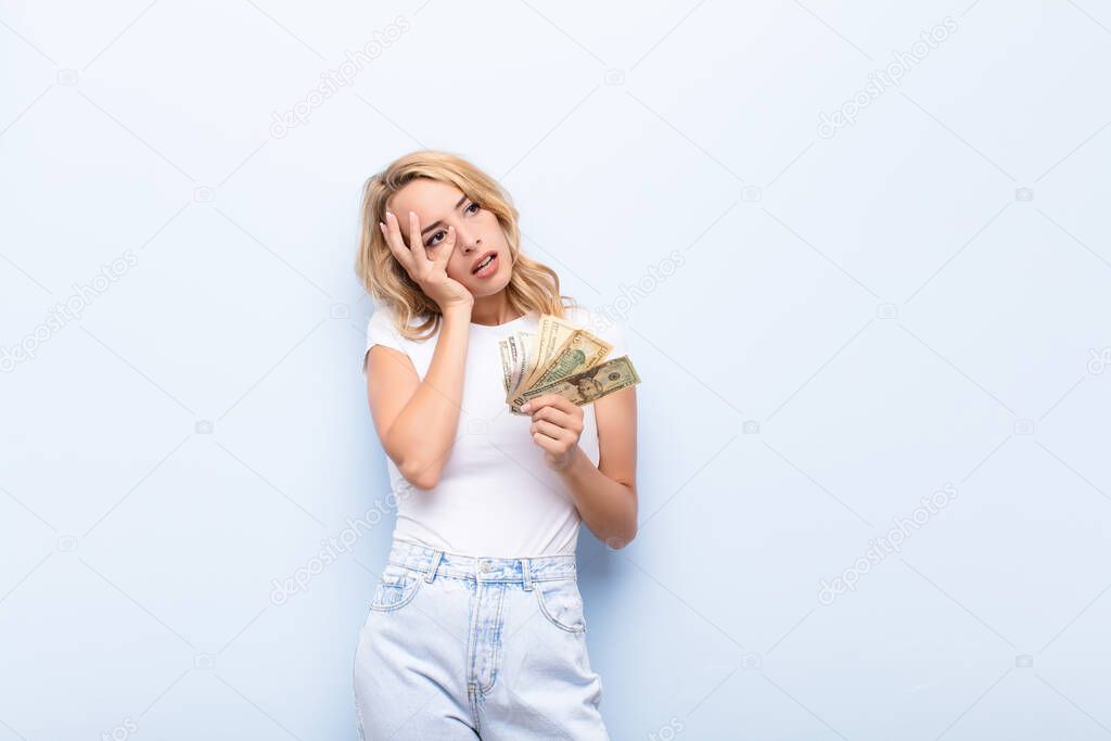 young blonde woman feeling bored, frustrated and sleepy after a tiresome, dull and tedious task, holding face with hand with dollar banknotes