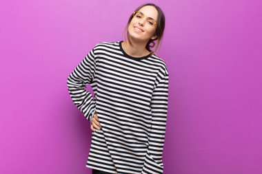 Genç ve güzel bir kadın neşeyle gülümsüyor, pozitif, mutlu, kendine güvenen ve rahatlamış bir ifadeyle mor duvara karşı.