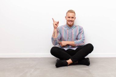 Genç sarışın adam mutlu bir şekilde gülümsüyor ve yan bakıyor, merak ediyor, düşünüyor ya da çimento zeminde oturuyor.