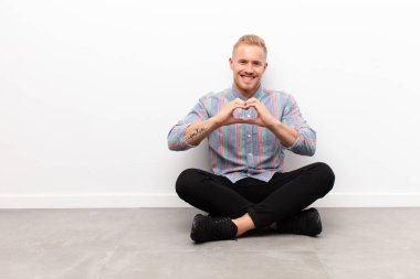 Genç sarışın adam gülümsüyor ve mutlu, sevimli, romantik ve aşık. İki elini de çimentoda oturtup kalp şeklinde yapıyor.