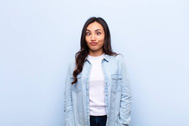 Aptal şaşı bir ifadeyle şapşal ve komik görünen genç Latin bir kadın, şaka yapıyor ve düz duvara karşı oynaşıyor.