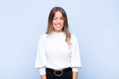 Genç ve güzel İspanyol kadın mutlu ve arkadaş canlısı görünüyor, gülümsüyor ve mavi duvara karşı pozitif bir tavırla sana göz kırpıyor.