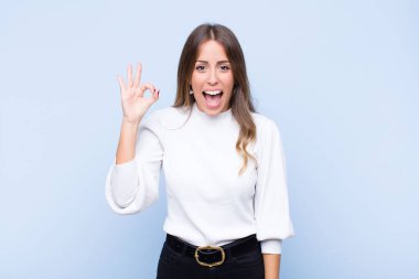 Genç ve güzel İspanyol kadın kendini başarılı ve tatmin olmuş hissediyor ağzı açık gülümsüyor, mavi duvara karşı elle işaret veriyor.