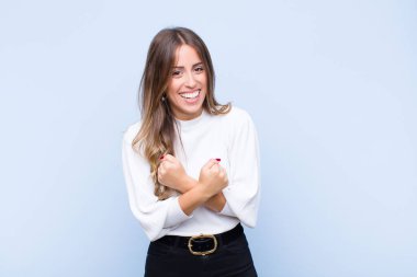 Genç ve güzel İspanyol kadın sevinçle gülümsüyor ve kollarını kavuşturup kollarını kavuşturuyor. Mavi duvara karşı mutlu ve pozitif hissediyor.