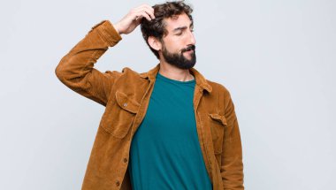 Genç yakışıklı adam şaşkın ve şaşkın hissediyor, başını kaşıyor ve düz duvara bakıyor.