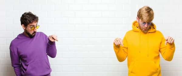 young cool men with open mouth pointing downwards with both hands, looking shocked, amazed and surprised against white tiles wall