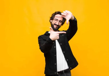 Genç çılgın yakışıklı adam mutlu, arkadaş canlısı ve pozitif hissediyor, gülümsüyor ve portre veya fotoğraf çerçevesi yapıyor. Elleri turuncu duvara dayalı.