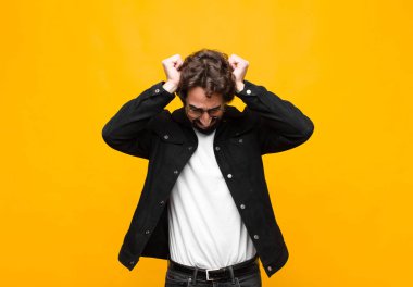 Genç çılgın yakışıklı adam stresli ve hayal kırıklığına uğramış hissediyor, el ele tutuşuyor, yorgun, mutsuz ve migreni turuncu duvara dayanmış.