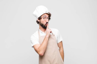 young crazy chef asking for silence and quiet, gesturing with finger in front of mouth, saying shh or keeping a secret against white wall clipart