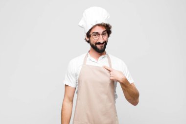 Genç çılgın şef mutlu, gururlu ve şaşırmış görünüyor, neşeli bir şekilde kendini gösteriyor, kendinden emin ve beyaz duvara karşı yüce hissediyor.