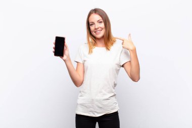 Kızıl saçlı, güzel bir kadın kendi gülüşünü işaret ediyor, pozitif, rahat, tatmin olmuş, akıllı bir telefon tutuyor.