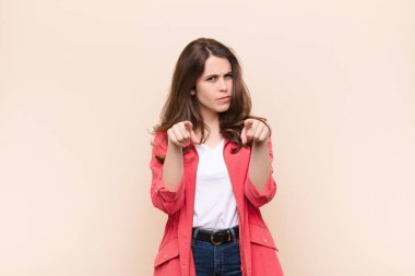 Genç ve güzel bir kadın kameraya hem parmaklarını hem de kızgın yüzünü göstererek bej arka plana karşı görevini yapmanı söylüyor.