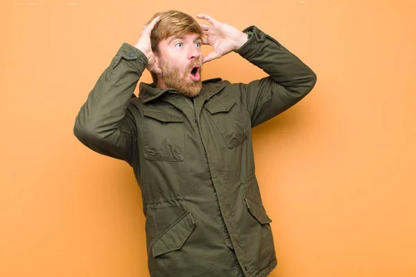 young blonde man with open mouth, looking horrified and shocked because of a terrible mistake, raising hands to head against flat wall