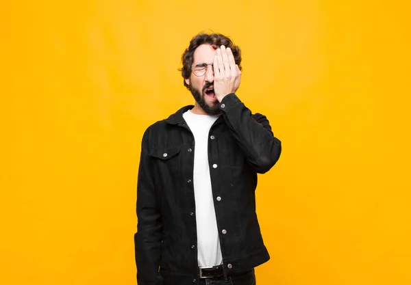 young crazy handsome man looking sleepy, bored and yawning, with a headache and one hand covering half the face against orange wall