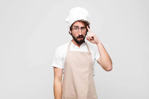 young crazy chef looking surprised, open-mouthed, shocked, realizing a new thought, idea or concept against white wall