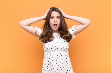Genç ve güzel bir kadın heyecanlı ve şaşırmış görünüyor, ağzı açık, iki eli başının üstünde, turuncu duvara karşı kendini şanslı hissediyor.