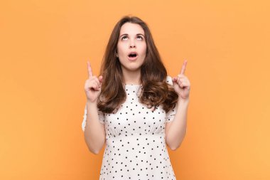 Genç ve güzel bir kadın huşu içinde ve ağzı açık bir şekilde turuncu duvara karşı şaşkın ve şaşkın bir bakışla yukarıya bakıyor.