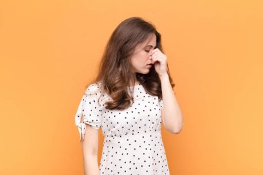 Genç ve güzel bir kadın stresli, mutsuz ve hayal kırıklığına uğramış, alnına dokunuyor ve turuncu duvara karşı şiddetli baş ağrısı çekiyor.