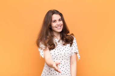 Genç, güzel bir kadın gülümsüyor, mutlu, kendinden emin ve arkadaş canlısı görünüyor, anlaşmayı bitirmek için el sıkışmayı teklif ediyor, turuncu duvara karşı işbirliği yapıyor.