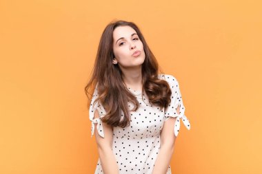 Genç, güzel bir kadın dudaklarını birbirine bastırıyor, tatlı, eğlenceli, mutlu, sevimli bir yüz ifadesiyle, turuncu duvara bir öpücük yolluyor.