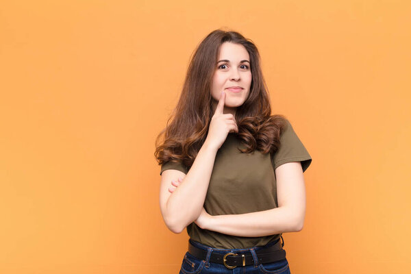 young pretty woman smiling with a happy, confident expression with hand on chin, wondering and looking to the side against orange wall