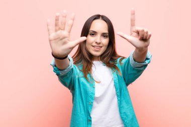 Genç, güzel bir kadın gülümsüyor ve arkadaş canlısı görünüyor. Elleri önde, pembe duvara doğru geri sayarak yedi ya da yedinci numarayı gösteriyor.