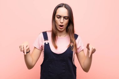 Genç, güzel bir kadın şoke olmuş, ağzı açık ve şaşırmış, pembe duvara bakıyor ve onu işaret ediyor.