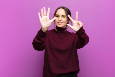 Genç, güzel bir kadın gülümsüyor ve arkadaş canlısı görünüyor. Sekiz ya da sekiz numarayı gösteriyor. Elleri önde, mor duvara doğru sayıyor.