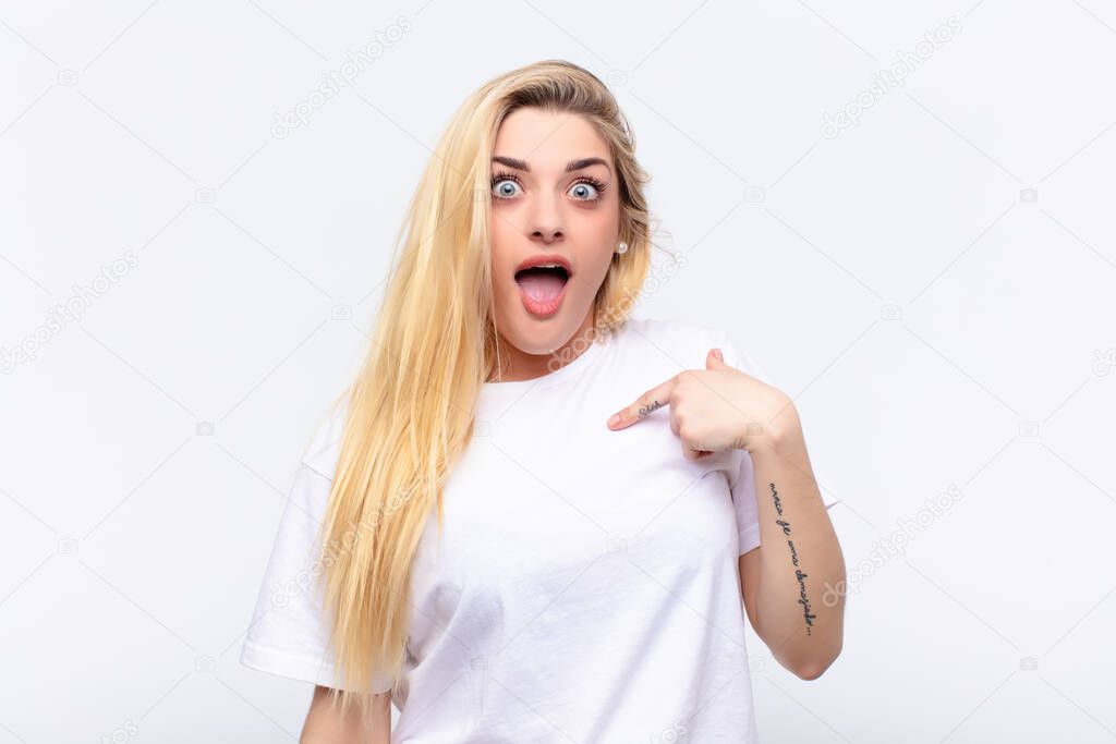 young pretty blonde woman looking happy, proud and surprised, cheerfully pointing to self, feeling confident and lofty against white wall