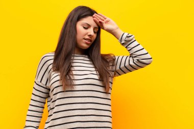 Genç ve güzel bir kadın stresli görünüyor, yorgun ve hayal kırıklığına uğramış, alnından ter damlıyor, umutsuz ve yorgun hissediyor sarı duvara karşı.