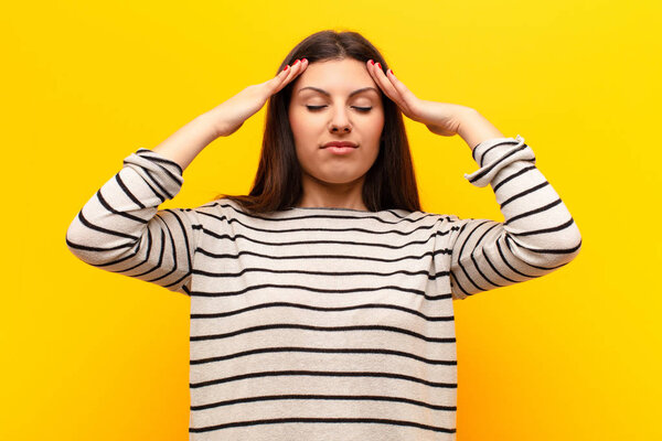 young pretty woman looking concentrated, thoughtful and inspired, brainstorming and imagining with hands on forehead against yellow wall