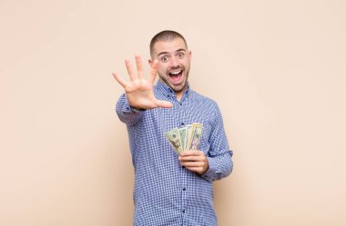 Genç, yakışıklı bir adam gülümsüyor ve arkadaş canlısı görünüyor. 5 ya da 5 numarayı gösteriyor. Elinde bir dolarla geriye doğru sayıyor.