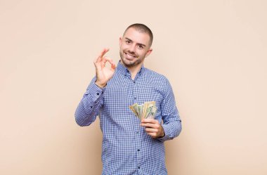 Genç yakışıklı adam kendini mutlu, rahat ve tatmin olmuş hissediyor. Onayını iyi bir hareketle gösteriyor. Dolar banknotlarıyla gülümsüyor.