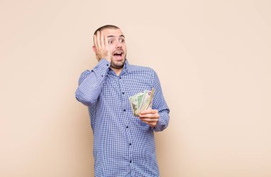 Genç yakışıklı adam mutlu, heyecanlı ve şaşırmış hissediyor, iki eli yüzü üzerinde bir dolar banknotuyla yan tarafa bakıyor.