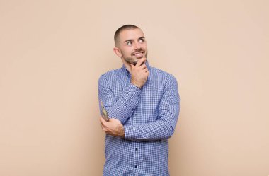 Genç yakışıklı adam mutlu bir şekilde gülümsüyor ve hayal kuruyor ya da şüphe ediyor, dolar banknotlarıyla yan yana bakıyor.