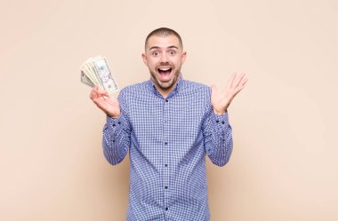 Genç yakışıklı adam mutlu, heyecanlı, şaşırmış ya da şaşırmış hissediyor, gülümsüyor ve şaşırıyor dolar banknotlarıyla inanılmaz bir şeye