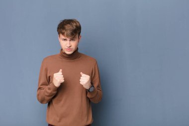 Genç sarışın adam kendinden emin, sinirli, güçlü ve agresif görünüyor. Yumrukları boks pozisyonunda dövüşmeye hazır. Düz duvara karşı izole.
