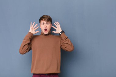 Genç sarışın adam ellerini havaya kaldırarak bağırıyor. Öfkeli, sinirli ve üzgün hissediyor. Duvara yaslanmış.