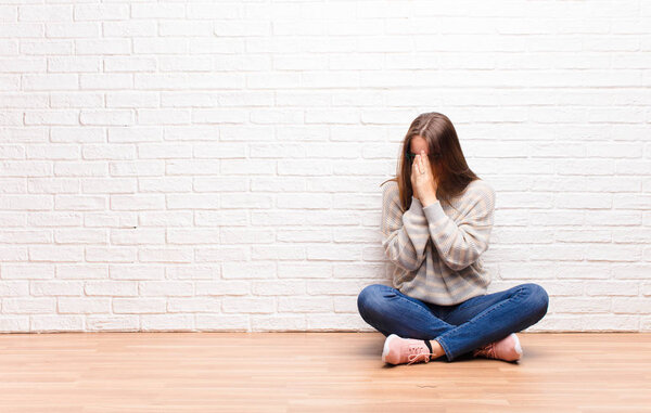 young blonde pretty girl covering eyes with hands with a sad, frustrated look of despair, crying, side view sitting on the floor