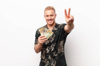 Genç sarışın adam gülümsüyor ve mutlu görünüyor, gamsız ve pozitif, bir eliyle beyaz duvara karşı zafer ya da barış işareti yapıyor.