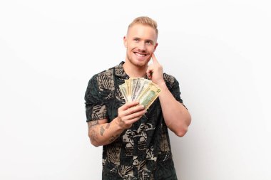 Genç sarışın adam mutlu bir şekilde gülümsüyor ve hayal kuruyor ya da şüpheyle beyaz duvara bakıyor.