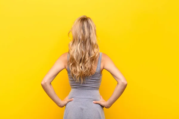年轻的金发女子感到困惑或满腹疑惑或疑问 双手叉腰靠着橙色的墙 想看看后面的风景 — 图库照片