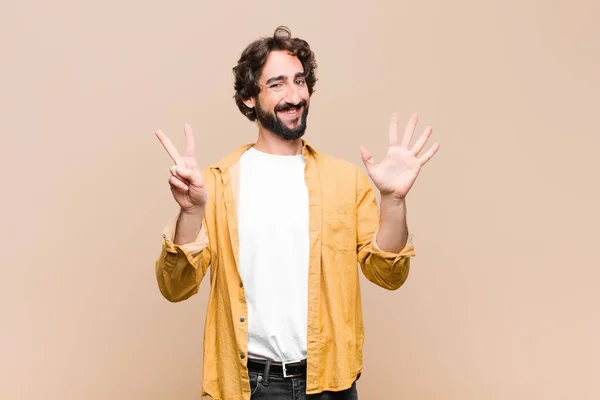 young crazy cool man smiling and looking friendly, showing number seven or seventh with hand forward, counting down against flat wall