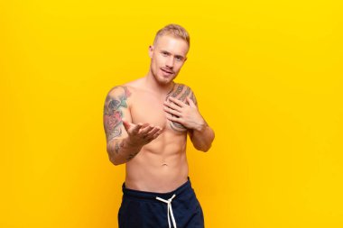 Genç güçlü sarışın adam mutlu ve aşık, bir eli kalbinin yanında gülümsüyor ve diğeri de sarı duvara yaslanmış.