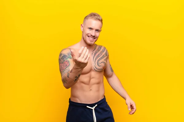 young strong blonde man feeling happy, successful and confident, facing a challenge and saying bring it on! or welcoming you against yellow wall