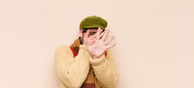 Genç, sakallı, sakallı, yüzü elle kaplayan ve diğer elini kamerayı durdurmak için öne koyan, fotoğraf ya da resimleri reddeden bir adam.