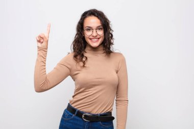 Genç, güzel bir kadın neşeyle ve mutlulukla gülümsüyor, tek eliyle yukarıya bakıyor ve beyaz duvara uzayı kopyalıyor.