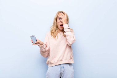 Genç sarışın kadın şok olmuş, korkmuş ya da korkmuş görünüyor. Yüzü elle kapatılmış ve akıllı telefon kullanarak parmaklarının arasından gizlice bakıyor.